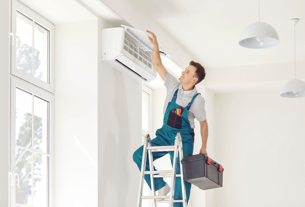 Electrician man standing on ladder holding toolbox maintaining air conditioner indoors.